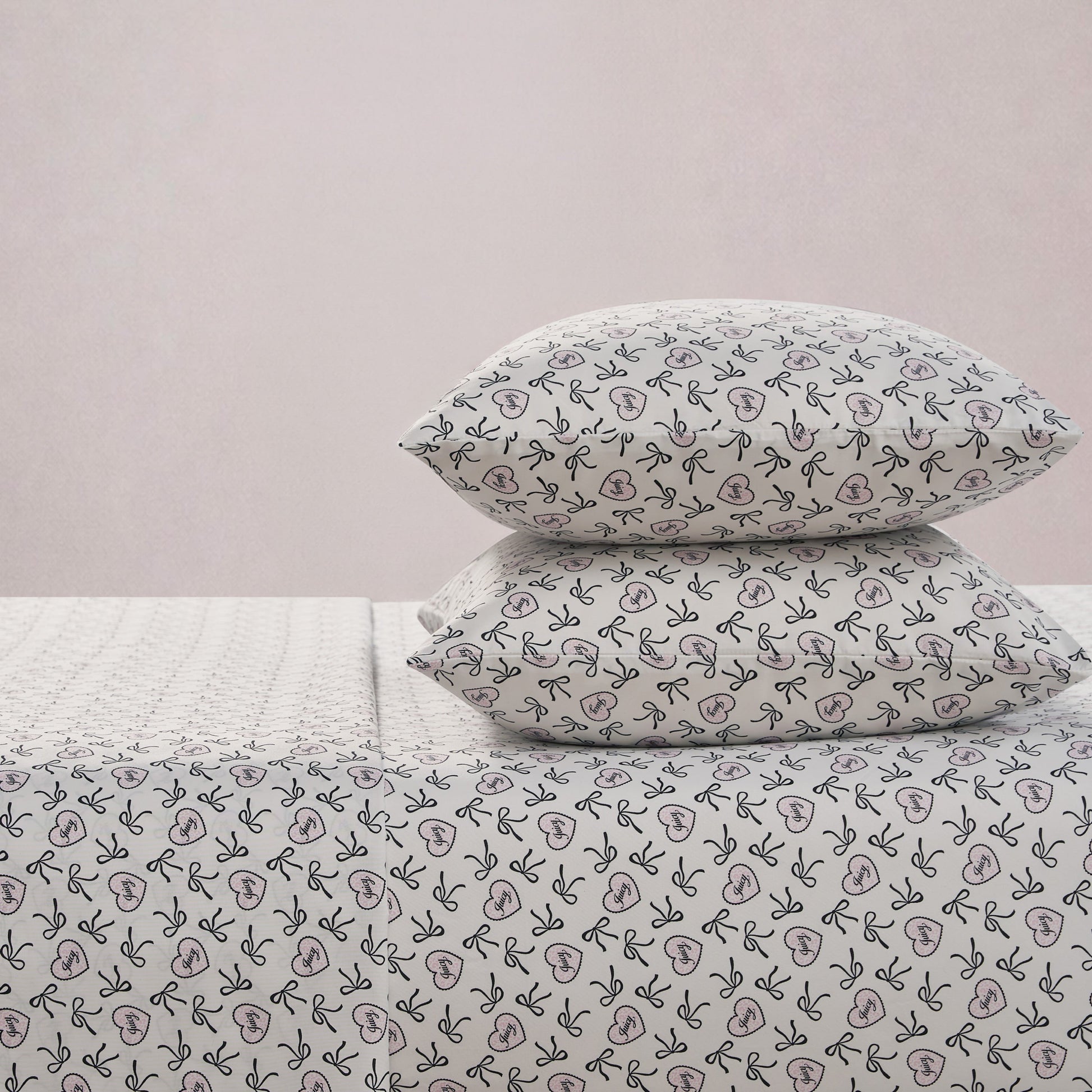 Two pillows stacked on a bed with matching white sheets, all decorated with simple black leaves and pink hearts. The Printed Microfiber Sheet Set adds a chic touch of comfort, set against a plain, light pink wall.
