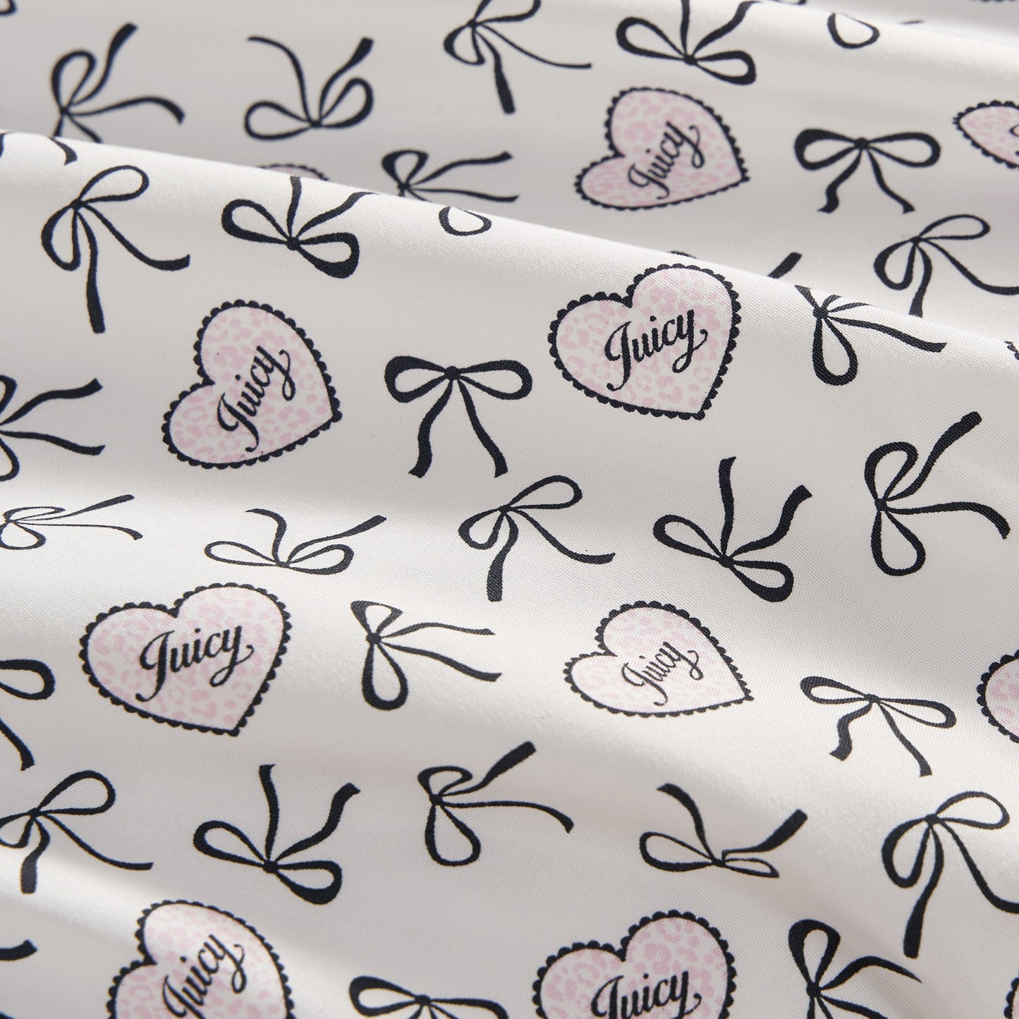 The Printed Microfiber Sheet Set features a white base with black bows and pink polka-dotted hearts outlined in black, each heart labeled "Juicy" in script. The luxury bedding is softly draped with gentle folds.