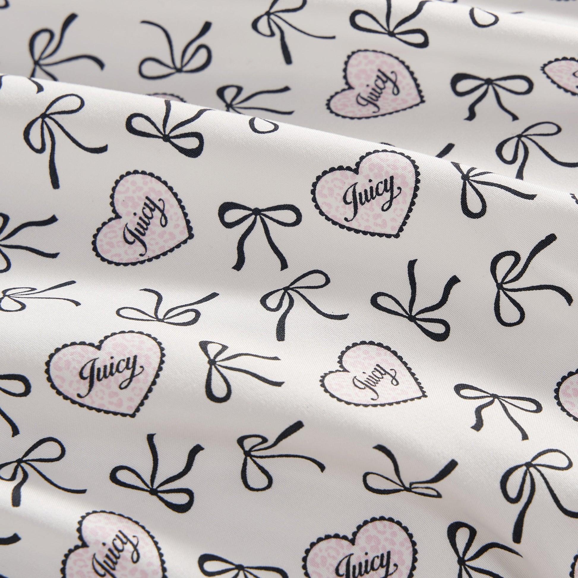 The Printed Microfiber Sheet Set features a white base with black bows and pink polka-dotted hearts outlined in black, each heart labeled "Juicy" in script. The luxury bedding is softly draped with gentle folds.