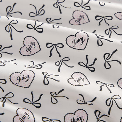The Printed Microfiber Sheet Set features a white base with black bows and pink polka-dotted hearts outlined in black, each heart labeled "Juicy" in script. The luxury bedding is softly draped with gentle folds.