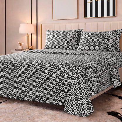 A neatly made bed features the Printed Microfiber Sheet Set in black and white with a repeating heart pattern, set in a modern bedroom with light walls, a lamp, and framed artwork above the bed.