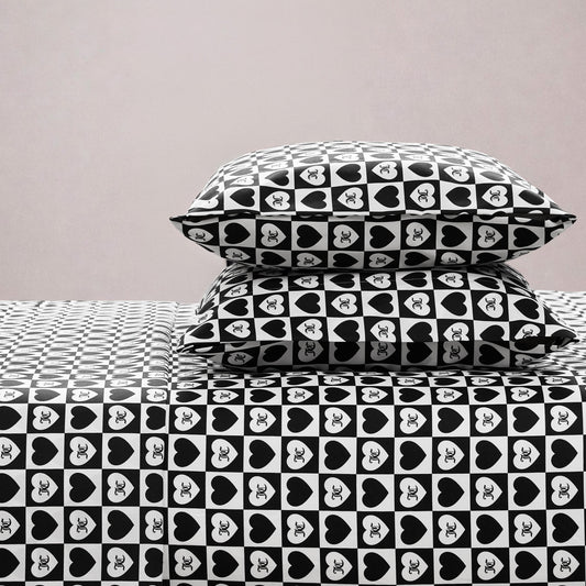 Two stacked pillows and a bed are dressed in the Printed Microfiber Sheet Set with black-and-white hearts featuring XO inside each heart, bringing stylish charm to your bedroom against a plain light-colored wall.