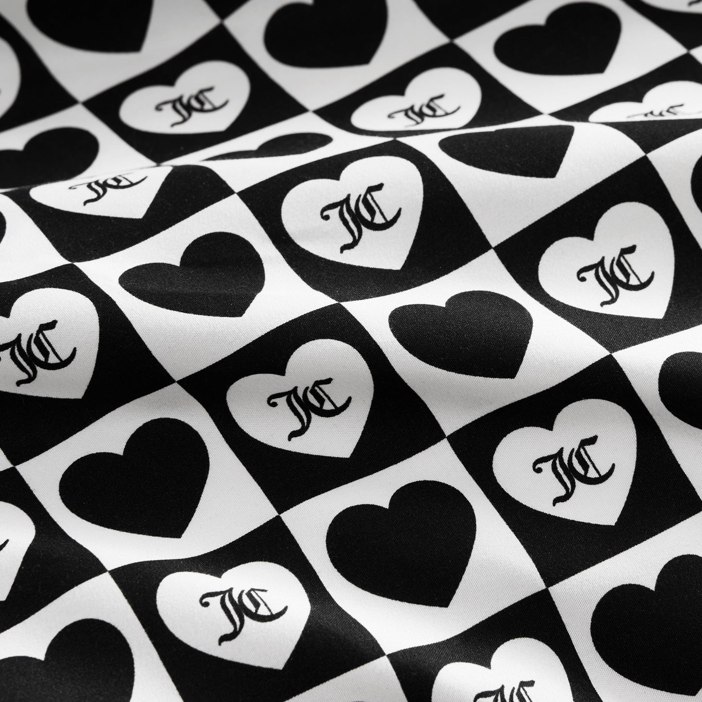 The Printed Microfiber Sheet Set features black and white checkerboard squares with alternating hearts, each displaying a stylized overlapping MC monogram. The luxury bedding surface has a subtle wavy texture.