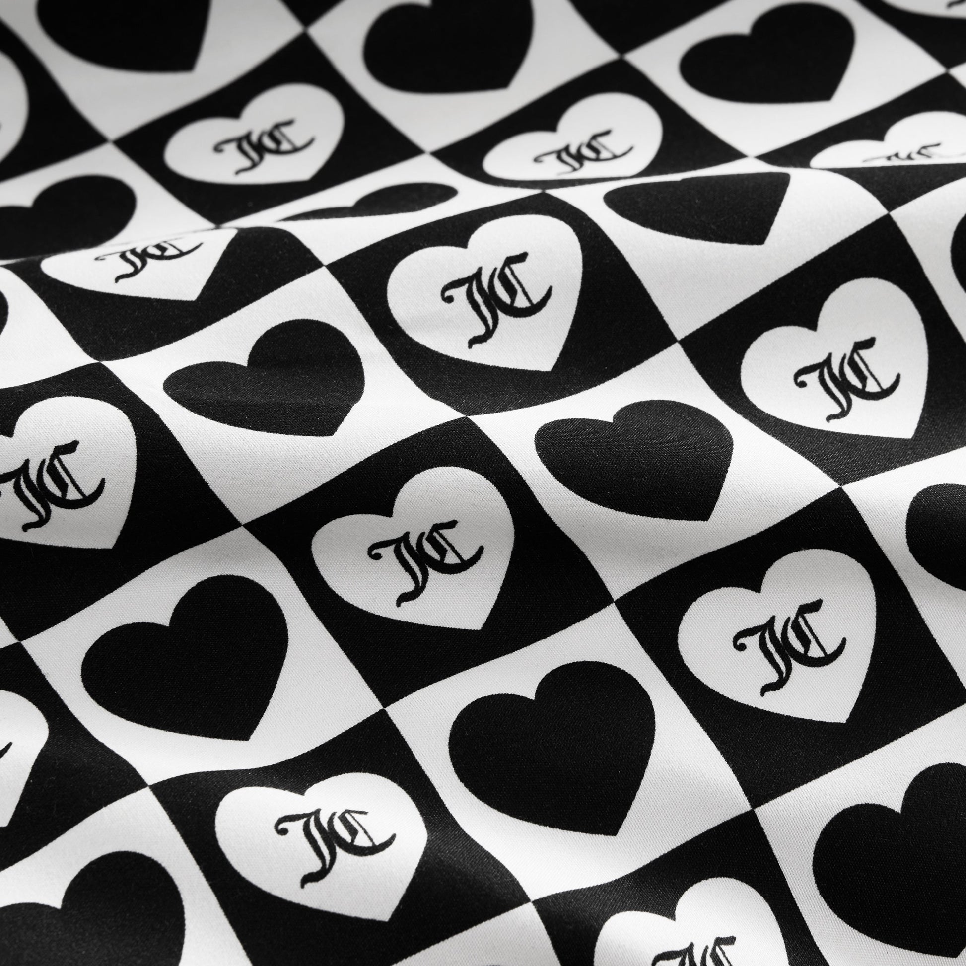 The Printed Microfiber Sheet Set features black and white checkerboard squares with alternating hearts, each displaying a stylized overlapping MC monogram. The luxury bedding surface has a subtle wavy texture.