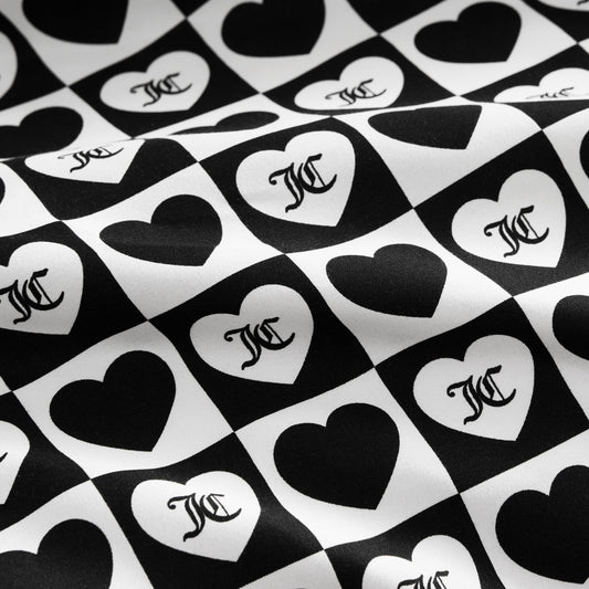 The Printed Microfiber Sheet Set features black and white checkerboard squares with alternating hearts, each displaying a stylized overlapping MC monogram. The luxury bedding surface has a subtle wavy texture.