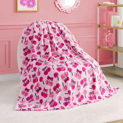 A Kids Plush Throw Blanket by Juicy Couture in soft pink with a vibrant cherry pattern is draped over a chair in a pink-themed room, with a bookshelf, wall art, and decorative mirror in the background.