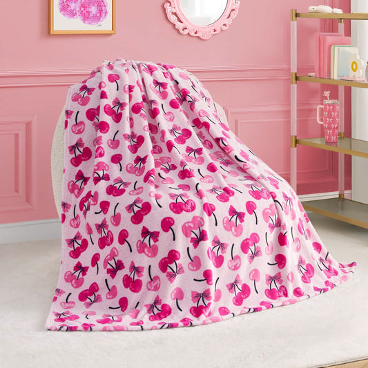 A Kids Plush Throw Blanket by Juicy Couture in soft pink with a vibrant cherry pattern is draped over a chair in a pink-themed room, with a bookshelf, wall art, and decorative mirror in the background.