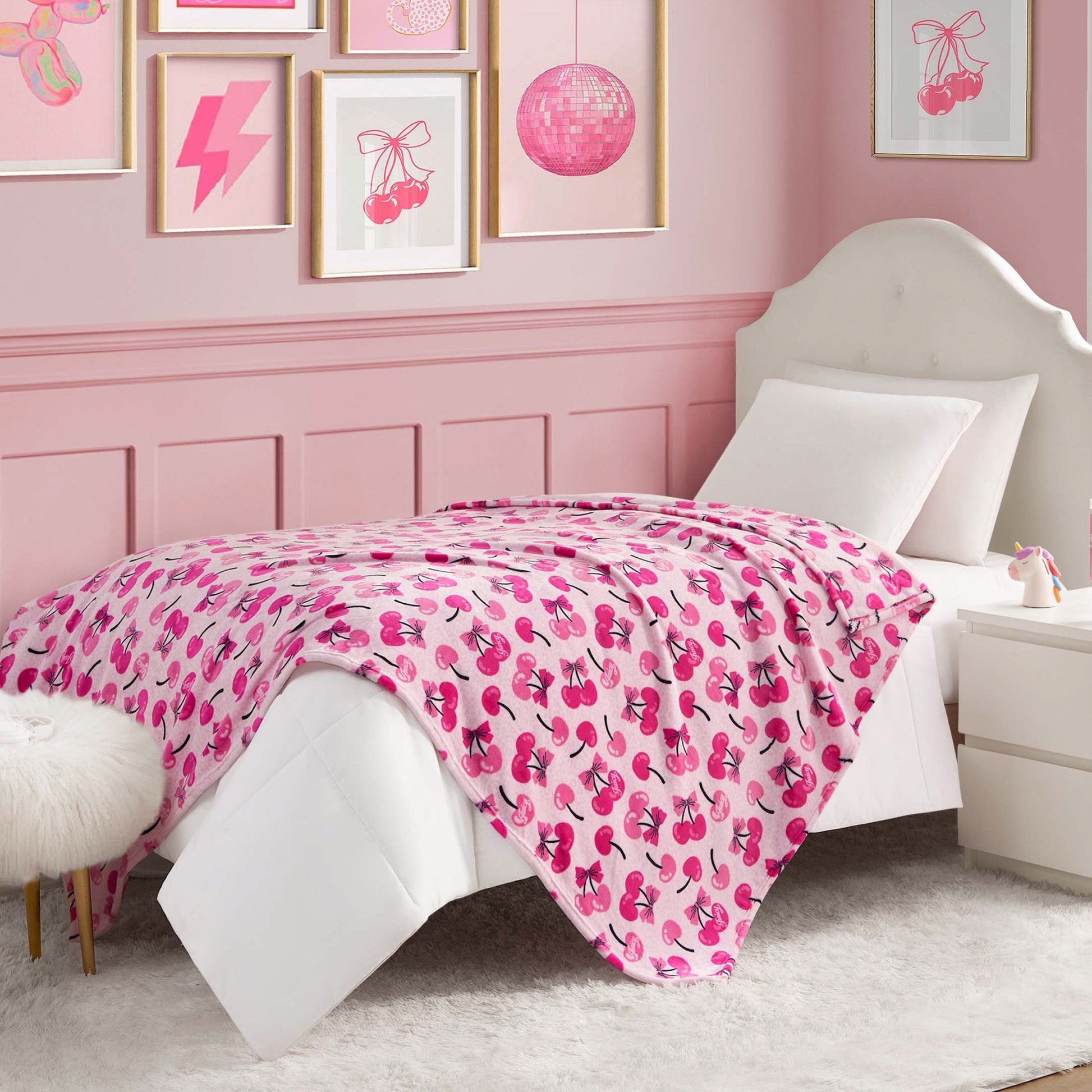 A white twin bed with a Kids Plush Throw Blanket in an ultra-soft pink-themed bedroom, featuring cherry and disco ball wall art, a nightstand, and a furry white stool.