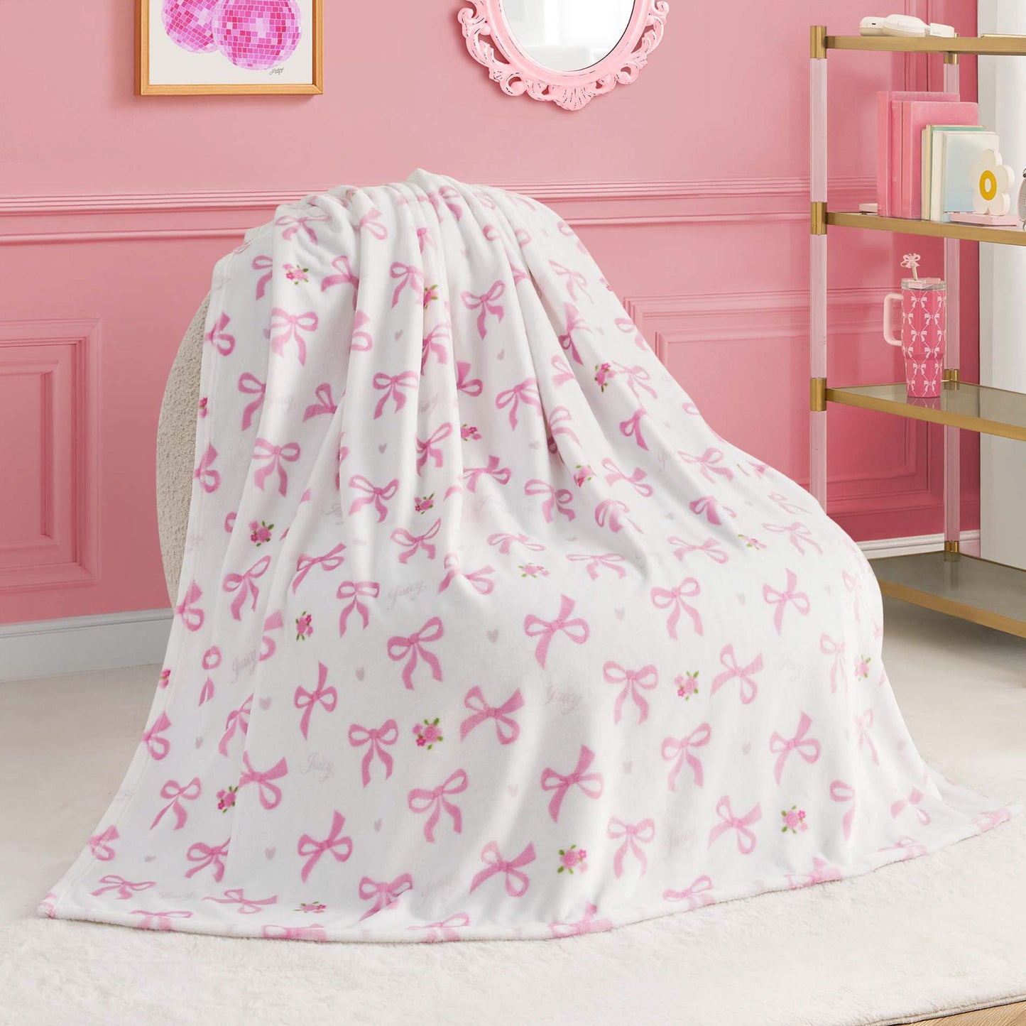 A Kids Plush Throw Blanket, ultra-soft and white with pink bows and small flower patterns, is draped over a chair in a pink-themed room featuring elegant wall molding, shelves, and framed art.