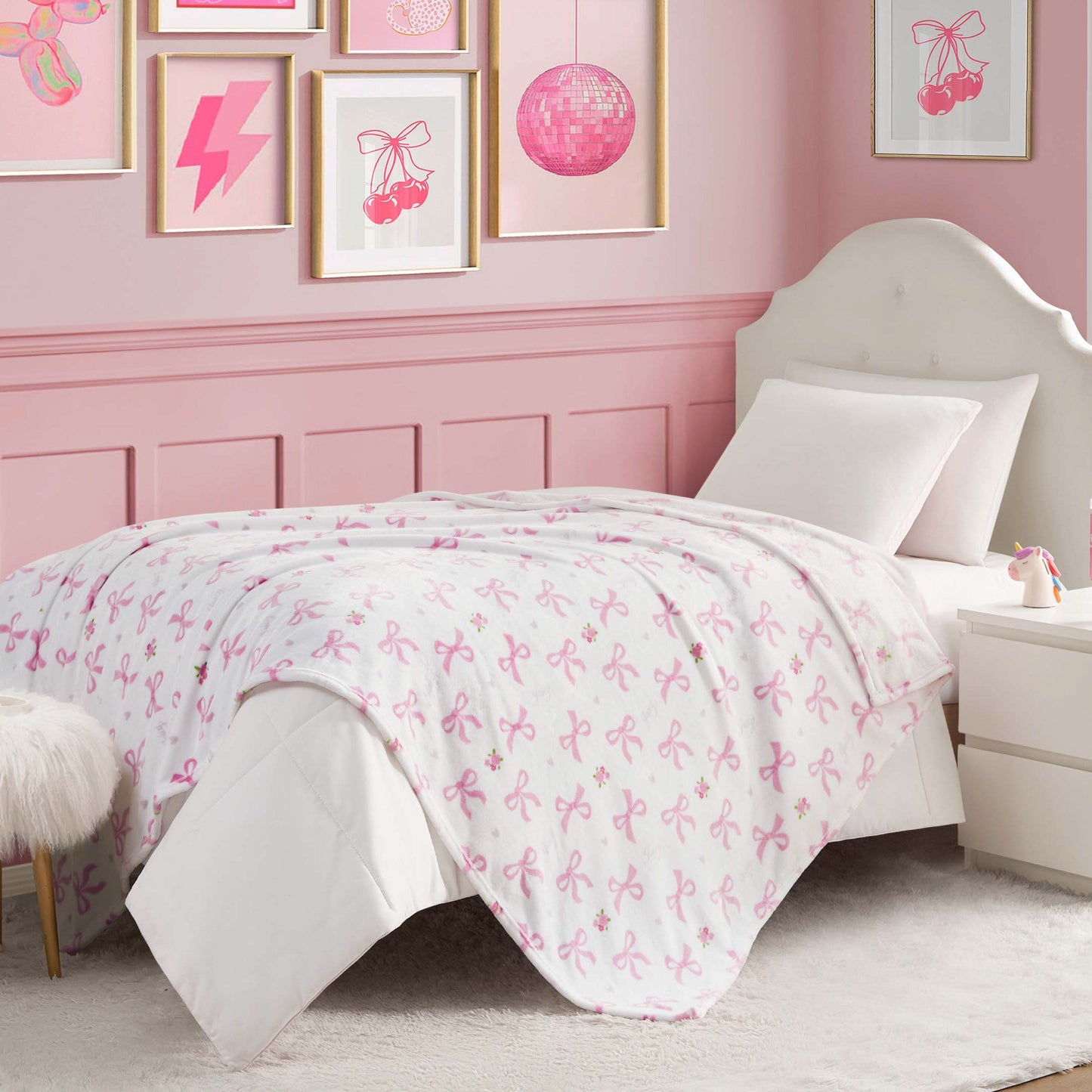 A neatly made bed with a Kids Plush Throw Blanket in a white and pink ribbon pattern, set in a pink-themed bedroom with framed art, a white upholstered headboard, and a white nightstand.