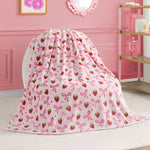 A Kids Plush Throw Blanket by Juicy Couture, featuring strawberries, pink bows, and "joy" in cursive, is draped over a white chair in a stylish pink room.