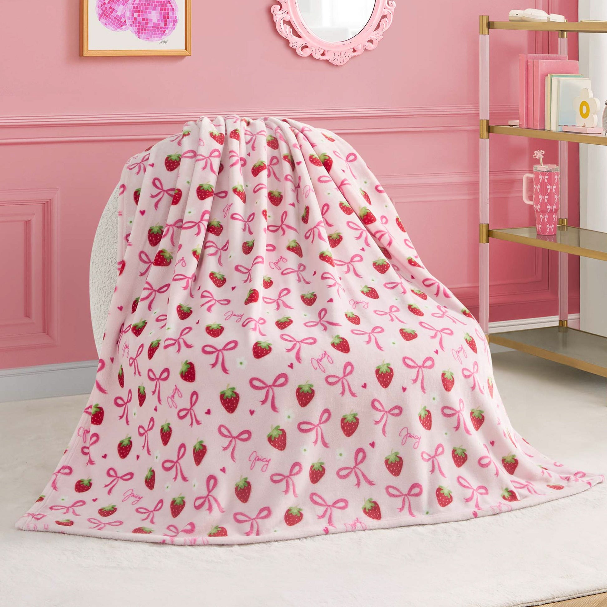 A Kids Plush Throw Blanket by Juicy Couture, featuring strawberries, pink bows, and "joy" in cursive, is draped over a white chair in a stylish pink room.