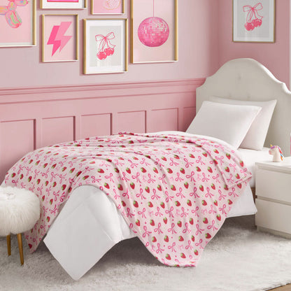 A pink-themed bedroom features a white bed topped with the Kids Plush Throw Blanket, decorated with pink ribbons. Pink and white wall art hangs above, with a white nightstand and furry stool for a cozy Juicy Couture Kids vibe.