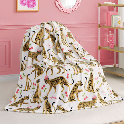 A Kids Plush Throw Blanket with a cheetah and pink flower design is draped over a chair in a stylish pink room decorated with wall art, a round mirror, and a gold shelving unit.
