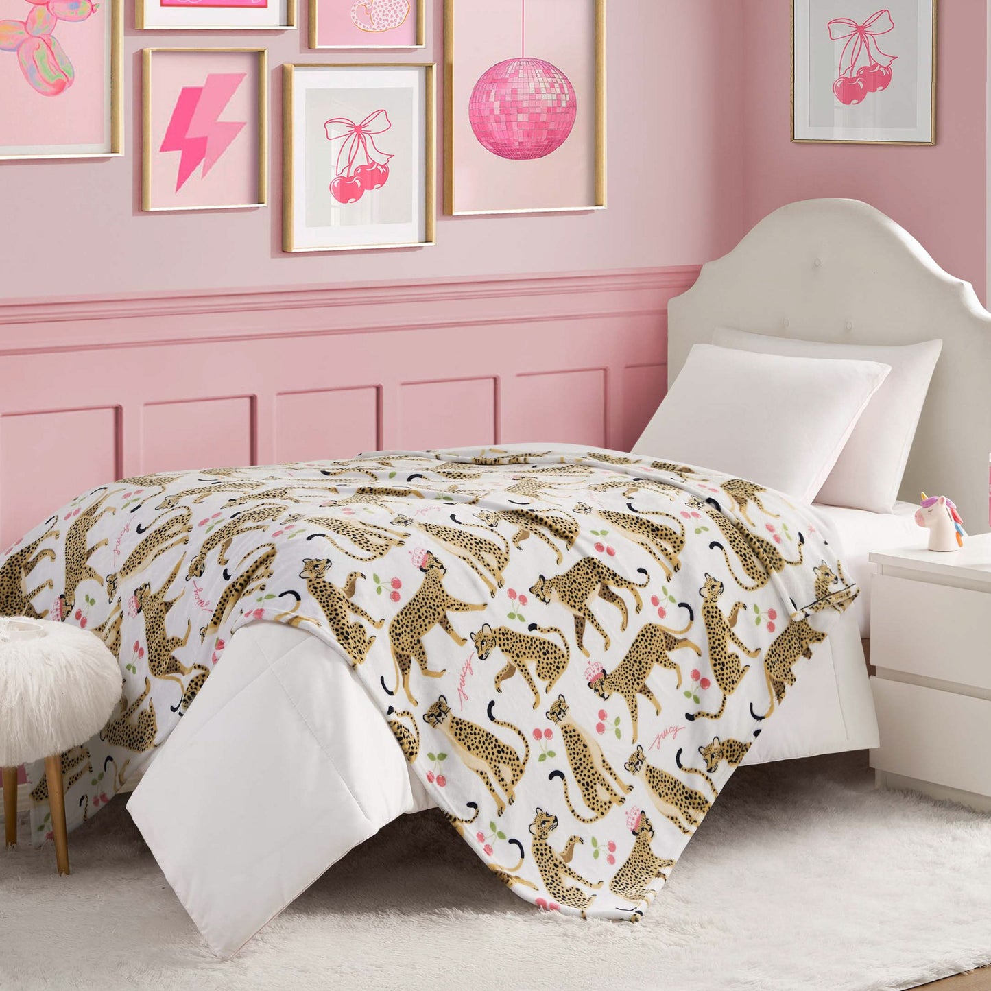 A pink-themed bedroom features a white headboard, framed art, a bedside table with lamp, and a fuzzy stool. The bed is neatly made with the Kids Plush Throw Blanket in white and leopard print for an ultra-soft touch.