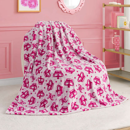 A Kids Plush Throw Blanket in pink and white with crown designs drapes over a chair in a stylish, pink-themed room with wall art, a mirror, and a gold shelving unit.