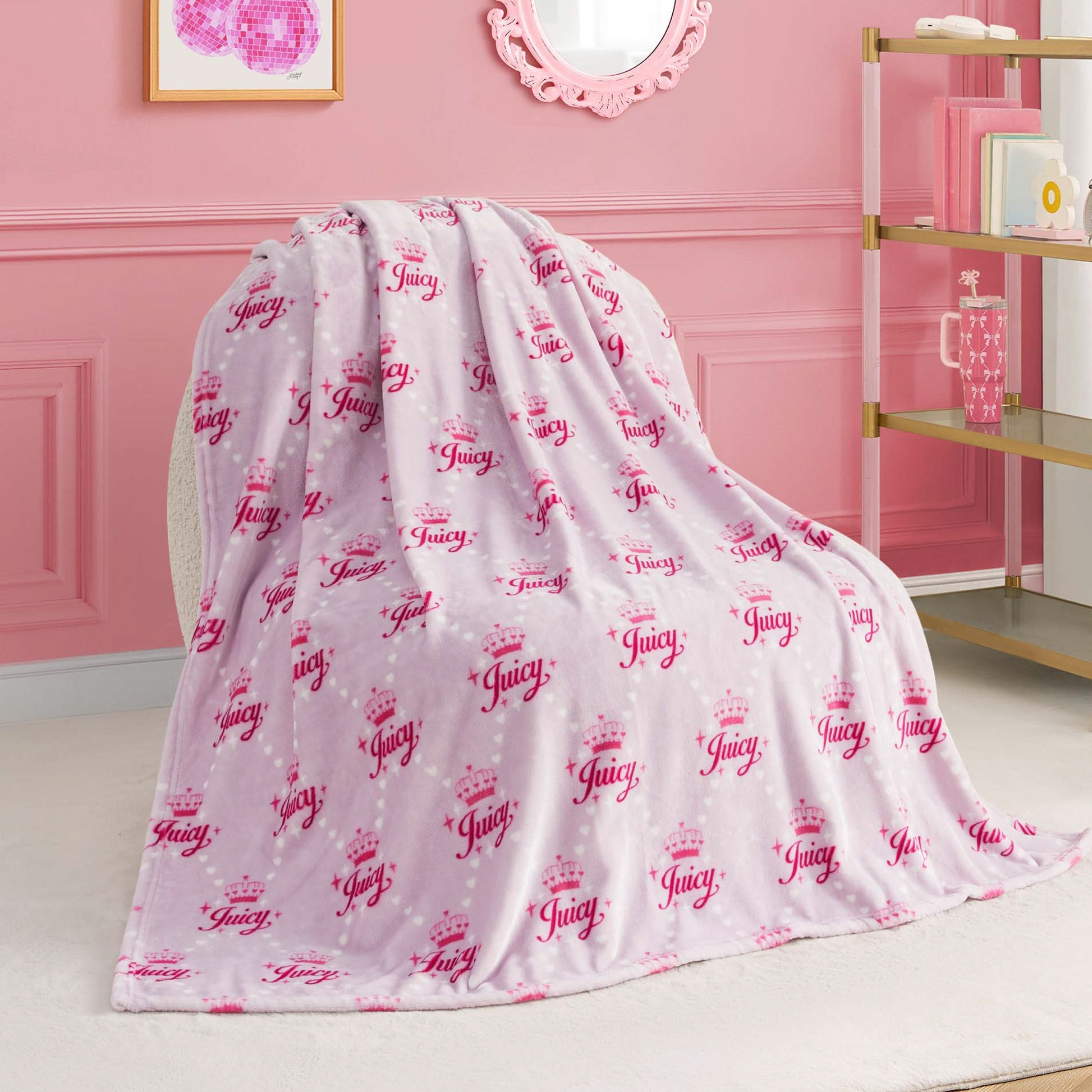 A Kids Plush Throw Blanket in pink and white with “Juicy” and crown designs is draped over a white chair in a chic pink-themed room with gold and white shelves.