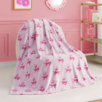 A Kids Plush Throw Blanket in pink and white with “Juicy” and crown designs is draped over a white chair in a chic pink-themed room with gold and white shelves.