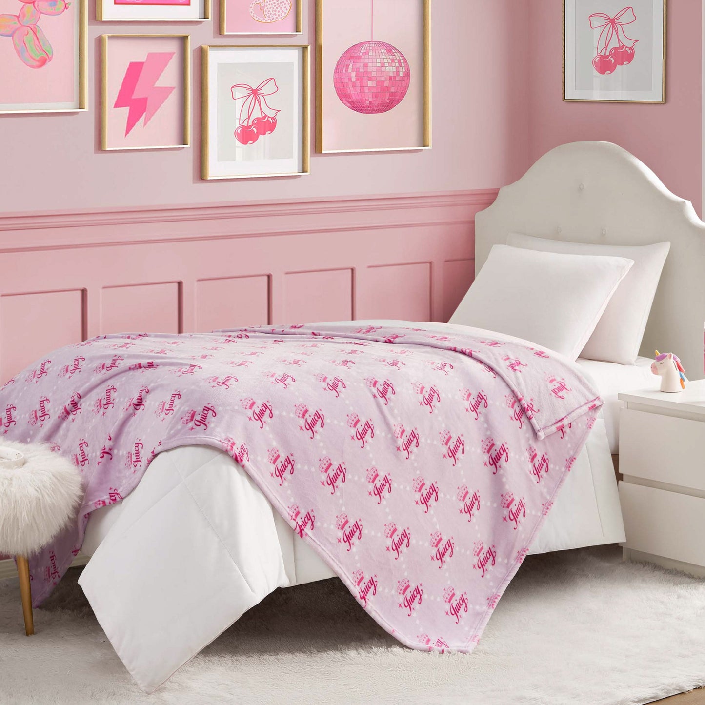 A neatly made bed with a white headboard and pink, white, and magenta bedding features the Kids Plush Throw Blanket for extra coziness. Pink-themed art, a white nightstand, and a furry stool complete the pink bedroom look.