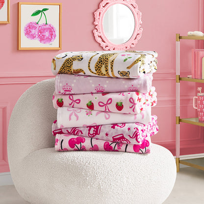 A stack of colorful Kids Plush Throw Blankets with playful patterns—leopards, crowns, strawberries, cherries—rests on a white textured chair in a pink room decorated with cherry artwork and a round mirror.