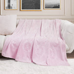 A Plush Oversized Throw Blanket, 60" x 90", in light pink with ornate embossed patterns is draped over a white couch beside a textured matching pillow, with black-and-white photos framed on the cream-colored wall behind.