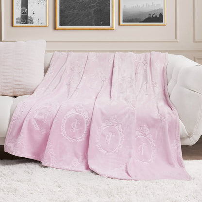 A Plush Oversized Throw Blanket, 60" x 90", in light pink with ornate embossed patterns is draped over a white couch beside a textured matching pillow, with black-and-white photos framed on the cream-colored wall behind.