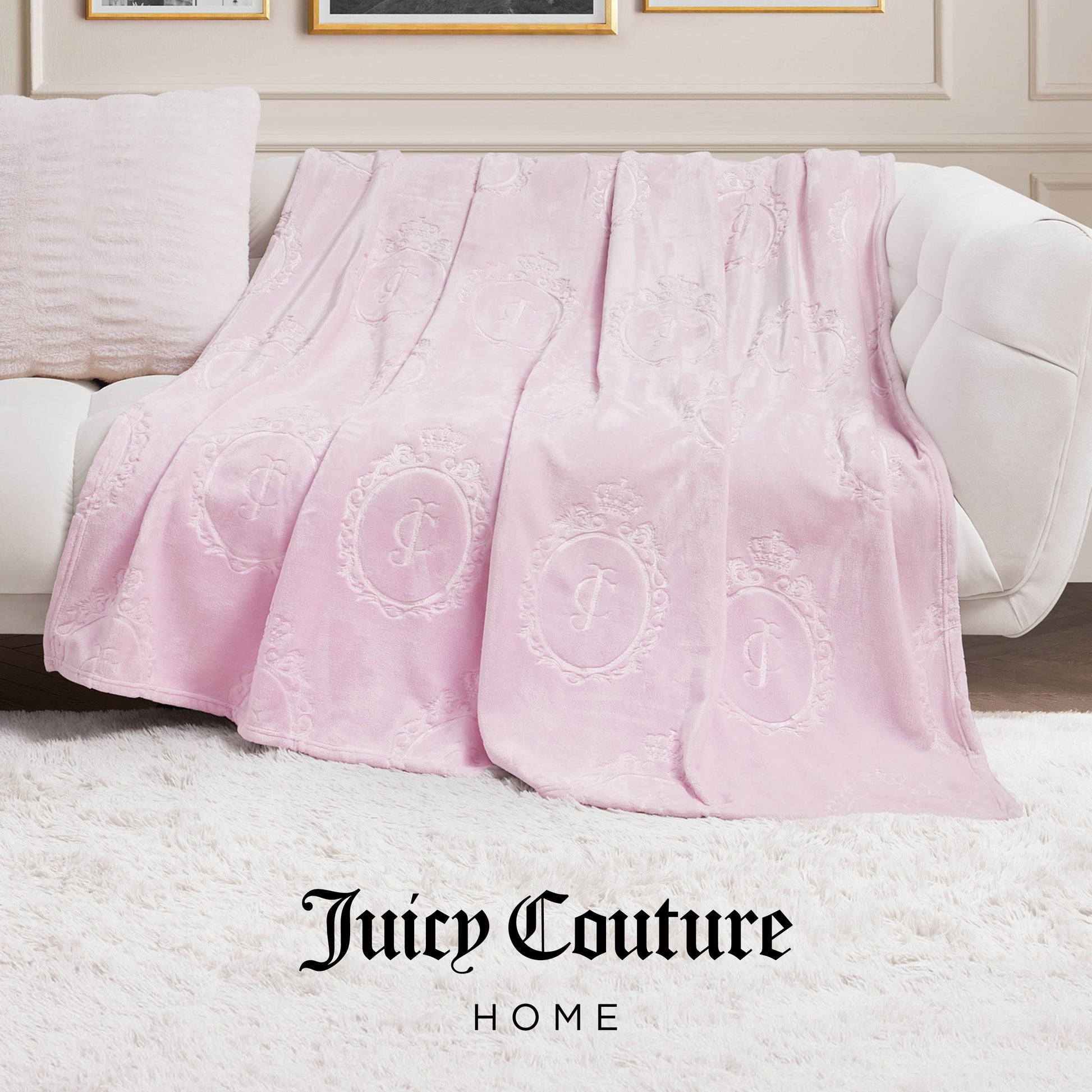 A light pink Plush Oversized Throw Blanket (60" x 90") with embossed logo patterns is draped over a white couch in a chic living room, featuring "Juicy Couture HOME" text at the bottom.