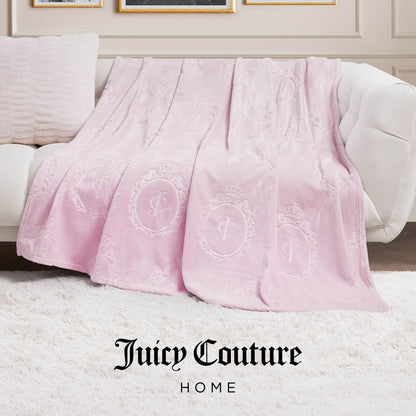 A light pink Plush Oversized Throw Blanket (60" x 90") with embossed logo patterns is draped over a white couch in a chic living room, featuring "Juicy Couture HOME" text at the bottom.