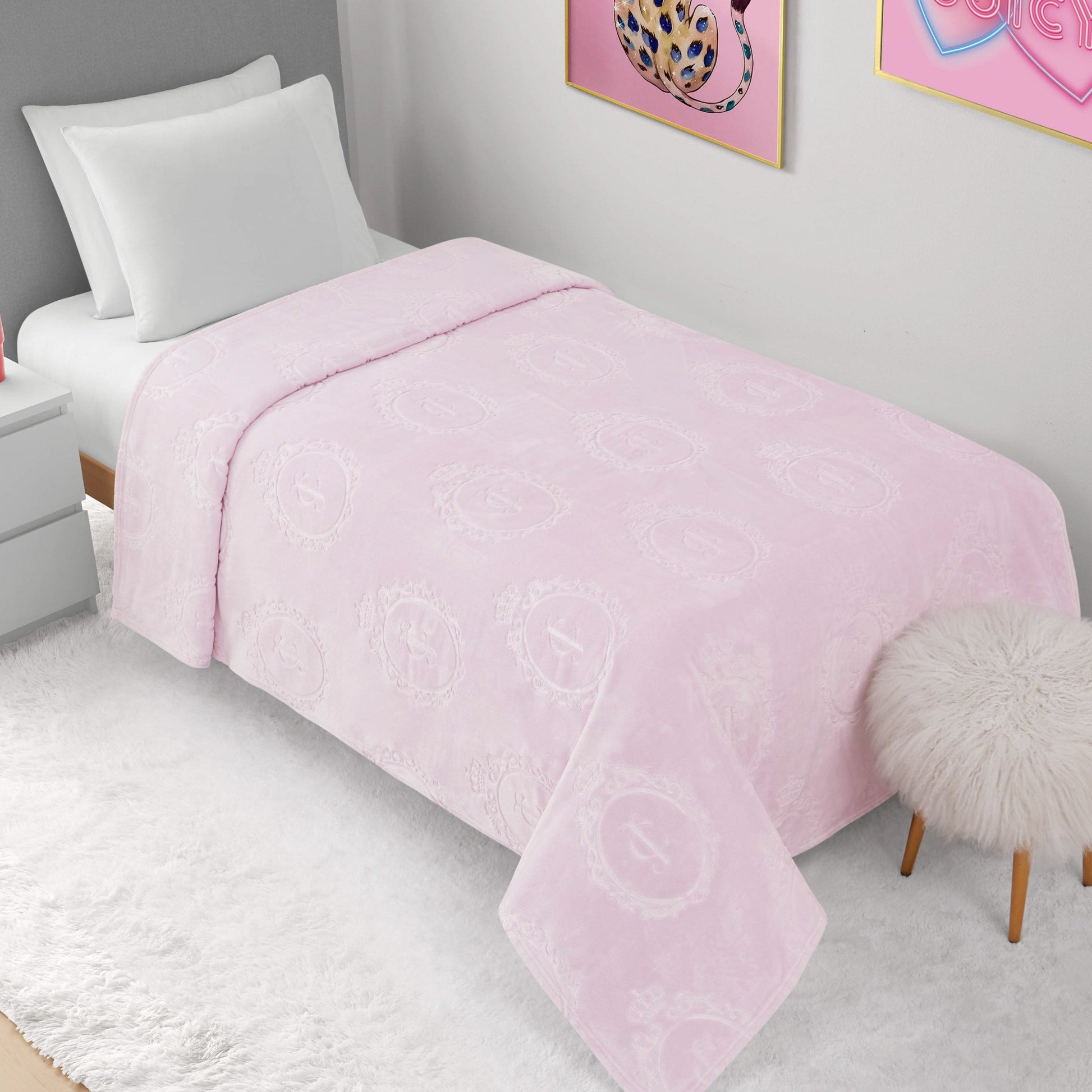 A neatly made bed features the Plush Oversized Throw Blanket (60" x 90") in soft light pink with subtle circular patterns, white pillows, white walls, pink wall art, a white nightstand, and a fuzzy stool on a white rug.