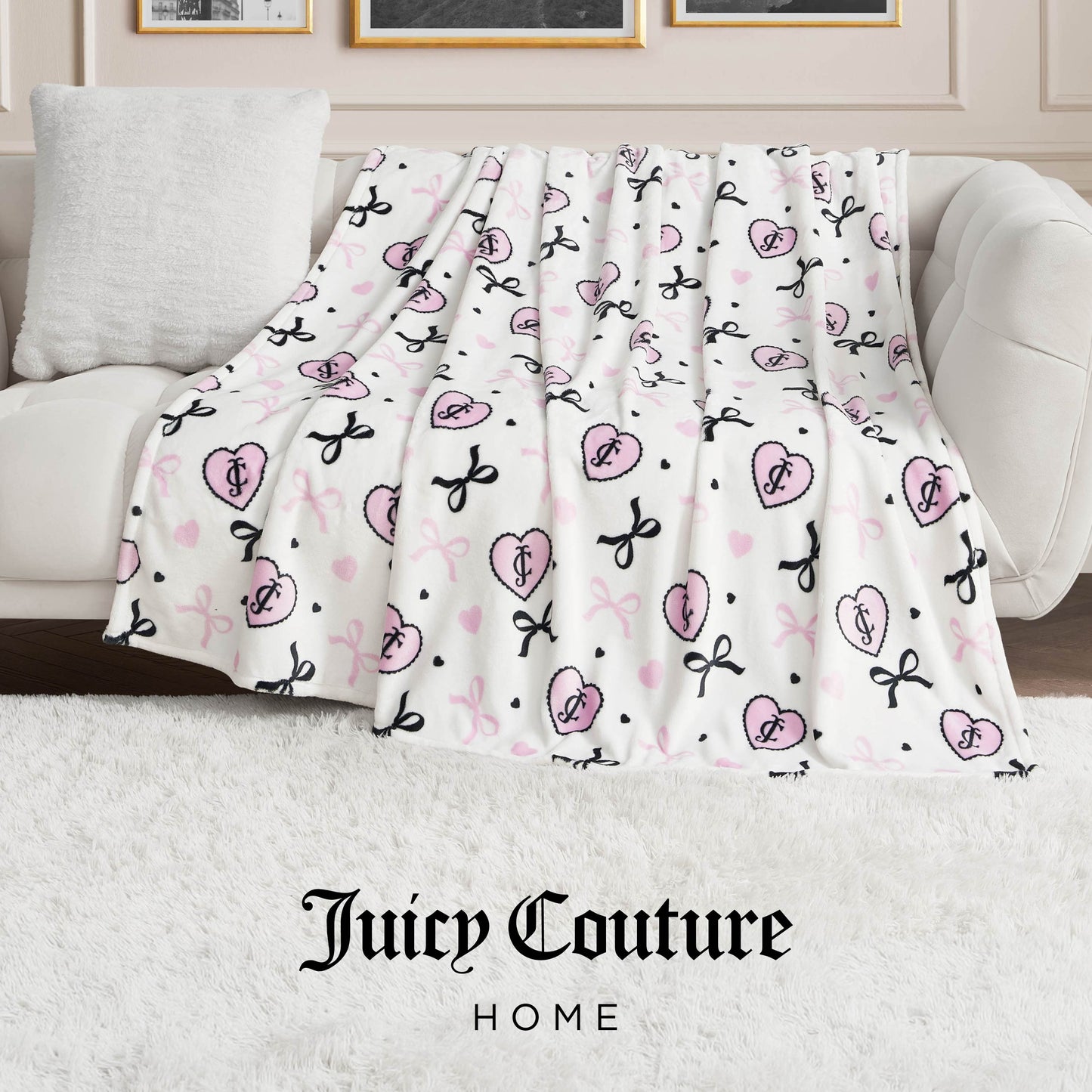 A white sofa with an ultra-soft Plush Oversized Throw Blanket, featuring pink and black Juicy Couture logos and bows, draped over the side. The scene includes a white pillow, plush white rug, and "Juicy Couture Home" text at the bottom.