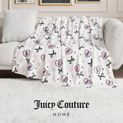 A white sofa with an ultra-soft Plush Oversized Throw Blanket, featuring pink and black Juicy Couture logos and bows, draped over the side. The scene includes a white pillow, plush white rug, and "Juicy Couture Home" text at the bottom.