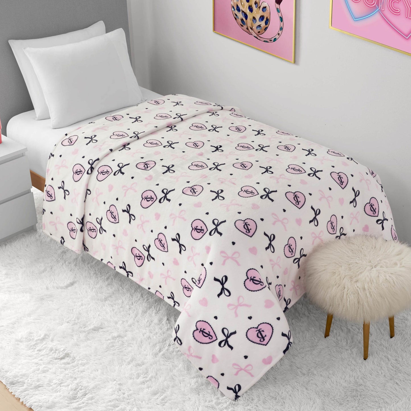 A neatly made twin bed with white bedding and a Plush Oversized Throw Blanket featuring bears, bows, and hearts. The room includes white walls, a fuzzy white rug, and a furry stool beside the bed.