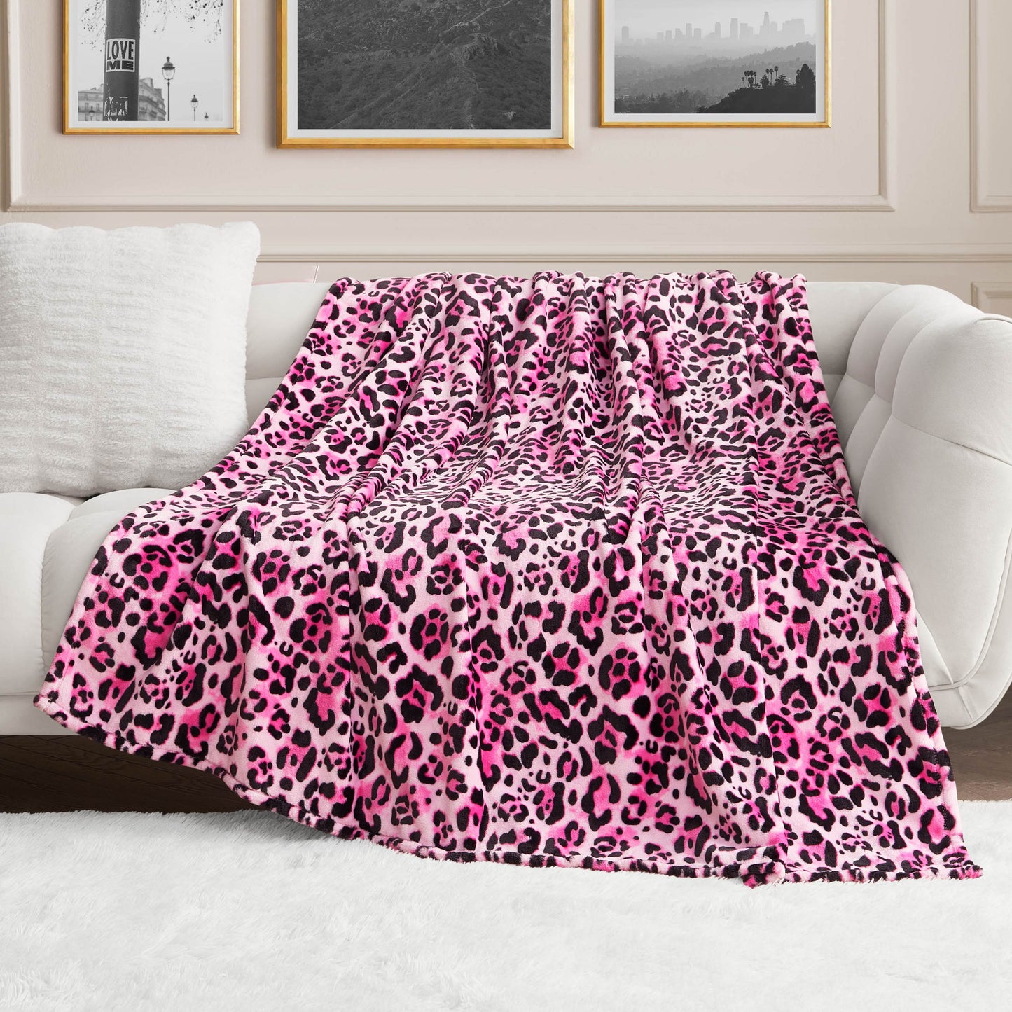 A stylish living room features a plush white sofa draped with the Plush Oversized Throw Blanket in pink and black leopard print, framed black-and-white photos on the wall, and a soft white rug on the floor.