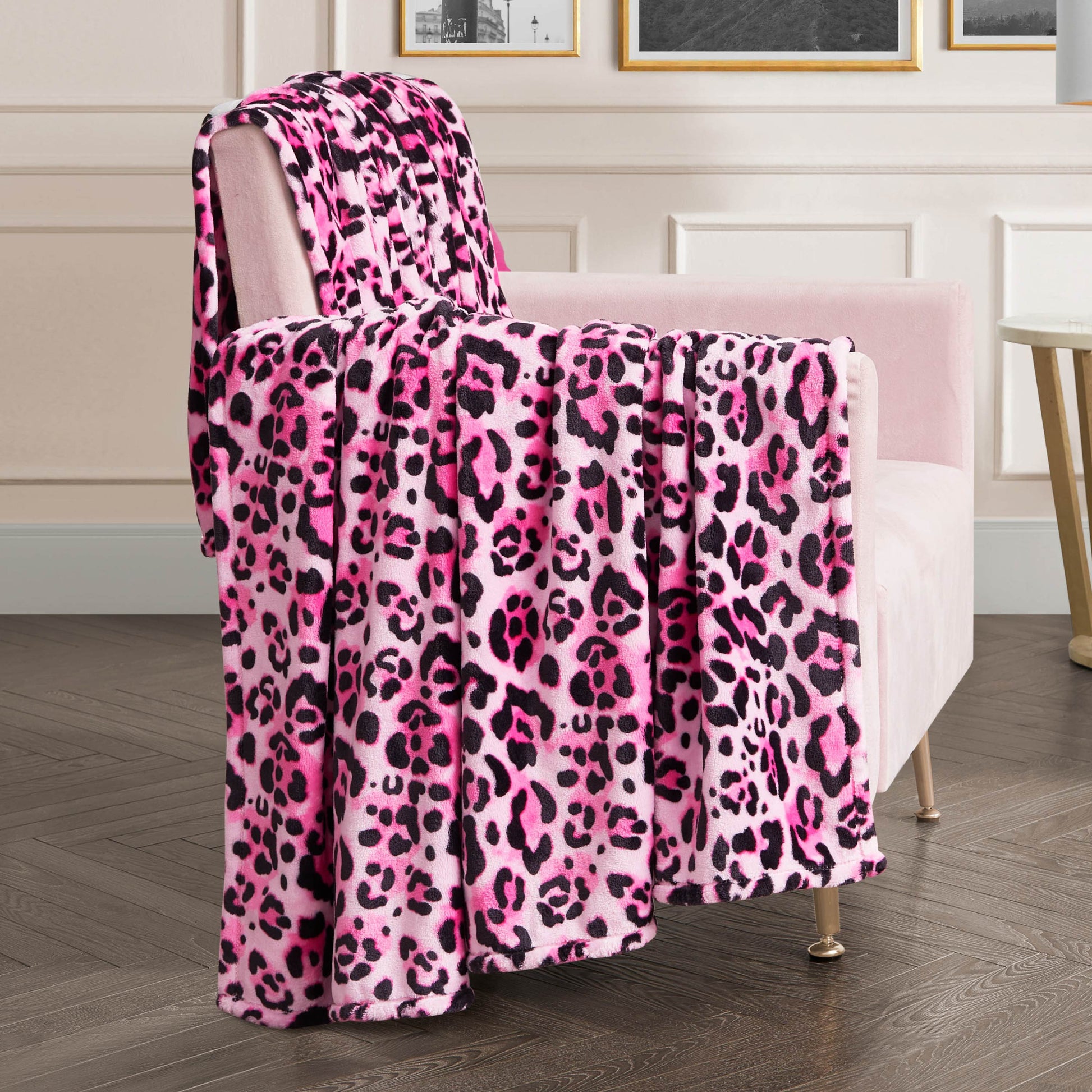 A stylish room with wood flooring and framed artwork features a plush pink chair draped with the Plush Oversized Throw Blanket in pink, black, and white leopard print over the back and seat.