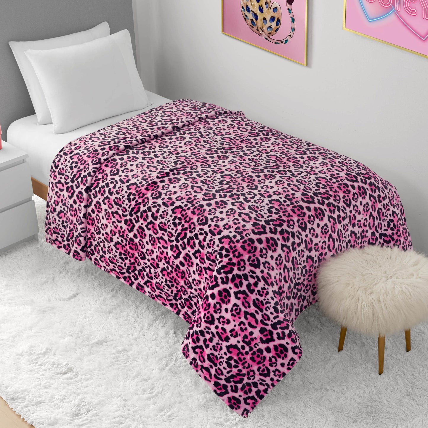 A neatly made bed with a Plush Oversized Throw Blanket in pink and black leopard print, white pillows, and a fluffy white rug. Nearby is a round furry stool and wall art with a pink heart and leopard illustration.
