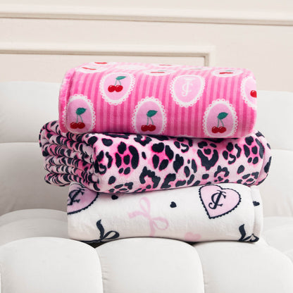 Three folded Plush Oversized Throw Blankets are stacked on a white tufted surface: the top is pink with cherries, the middle has a pink and black leopard print, and the bottom features white with pink and black hearts and bows.