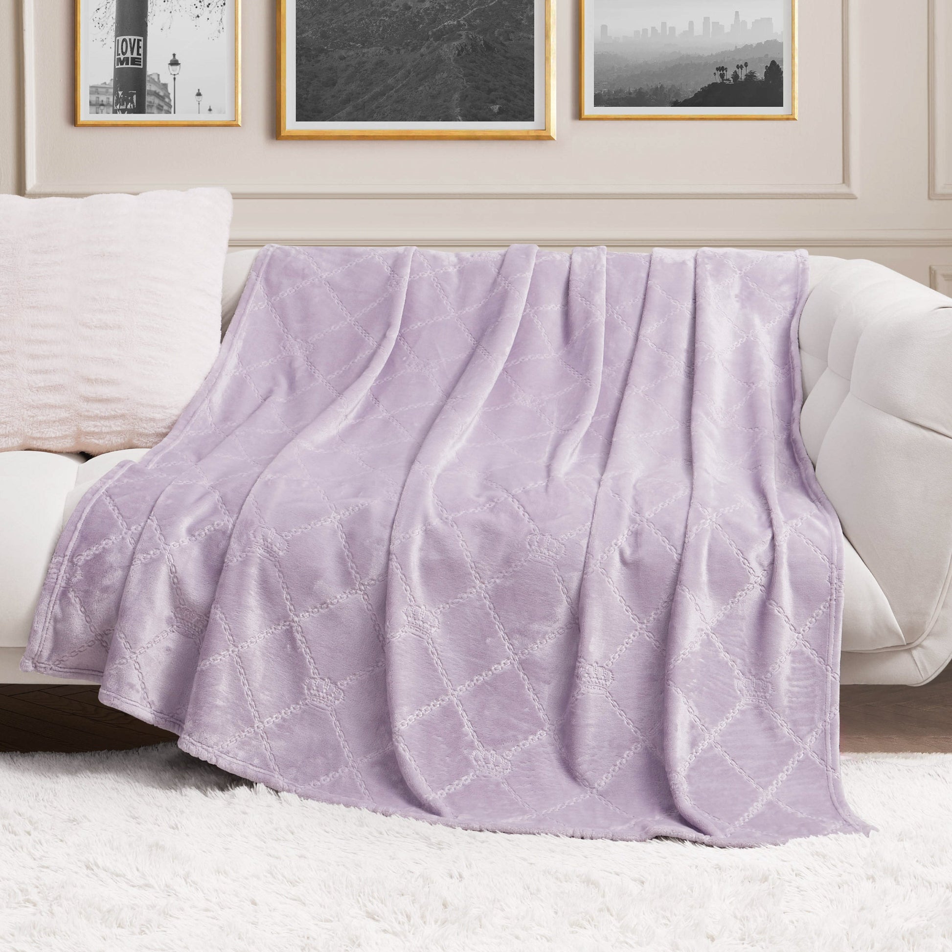A Plush Oversized Throw Blanket, 60" x 90", in soft lavender with a subtle pattern is draped over a white sofa in a chic living room featuring framed art and a fluffy white rug.