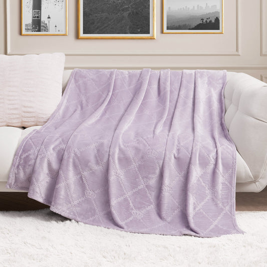 A Plush Oversized Throw Blanket, 60" x 90", in soft lavender with a subtle pattern is draped over a white sofa in a chic living room featuring framed art and a fluffy white rug.