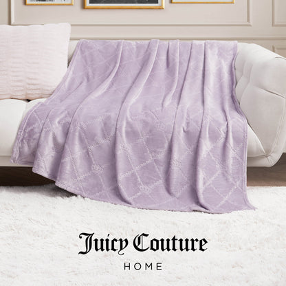 A light purple Plush Oversized Throw Blanket (60" x 90") with a quilted pattern is draped over a beige sofa next to a cream pillow on a white shag rug, with Juicy Couture Home at the bottom of the image.