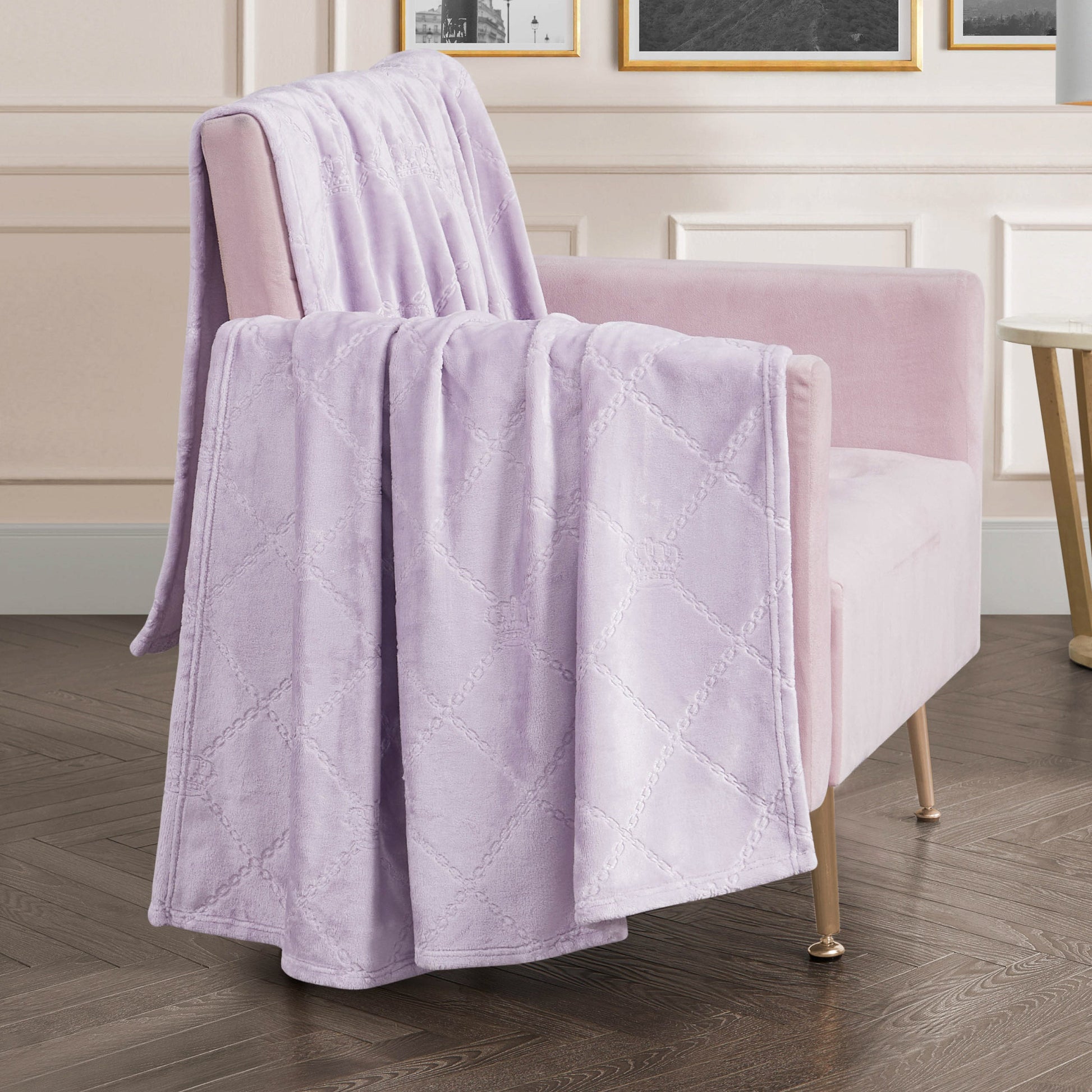 A Plush Oversized Throw Blanket, 60" x 90", in soft lavender with a diamond pattern is draped over a light pink velvet armchair in a chic living room with wood flooring and white paneled walls.