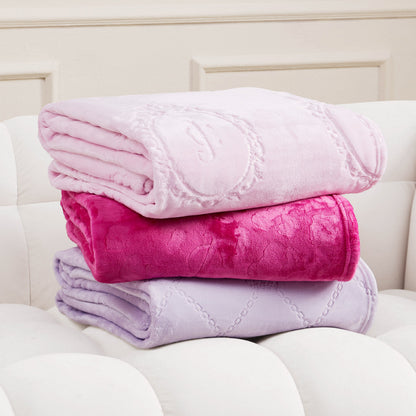 Three soft, folded Plush Oversized Throw Blankets (60" x 90") in pastel pink, bright pink, and lavender are stacked on a white tufted couch. The top blanket features quilted patterns and an embroidered monogram.