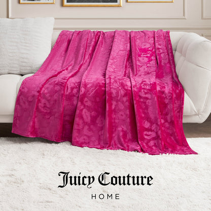 A vibrant pink Plush Oversized Throw Blanket, 60" x 90", is draped over a light sofa in a chic living room with a white rug and classic wall moulding. The Juicy Couture Home logo appears at the bottom of the blanket.