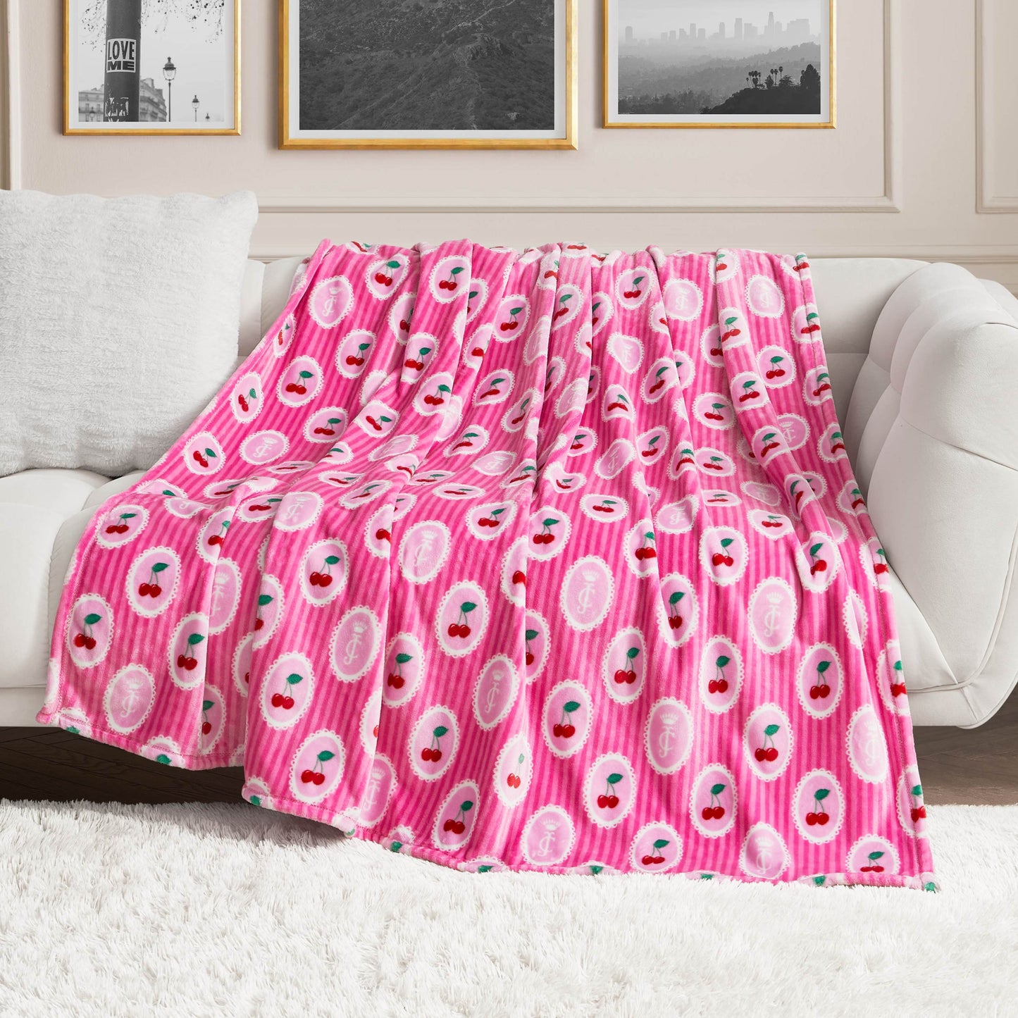 A Plush Oversized Throw Blanket in pink with smiling cherry design is draped over a white couch in a modern living room featuring framed photos and a fuzzy white rug.