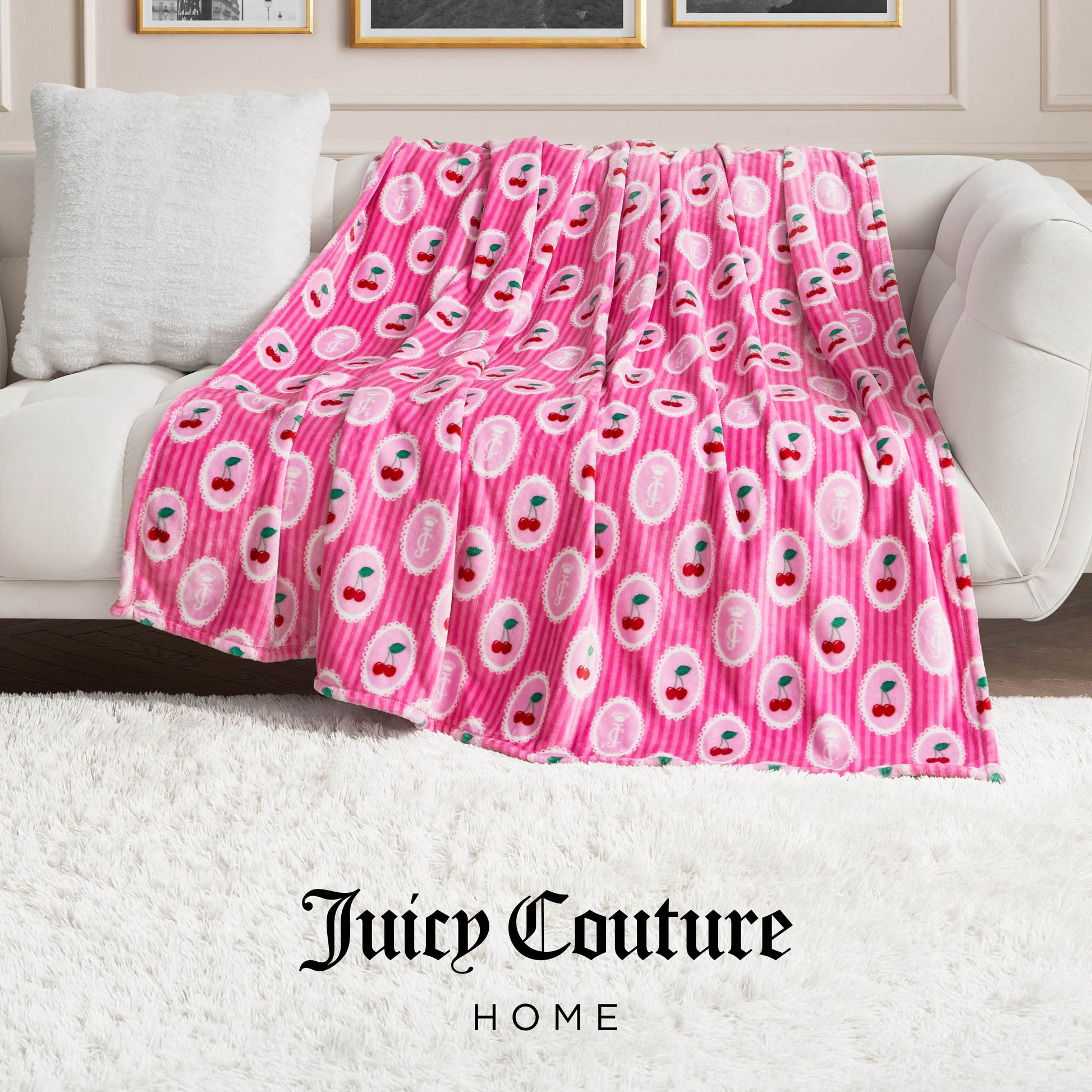 A Plush Oversized Throw Blanket in pink with cherry and Juicy Couture patterns is draped over a white sofa in a stylish living room with a white rug and elegant wall paneling. The Juicy Couture Home logo is visible at the bottom.