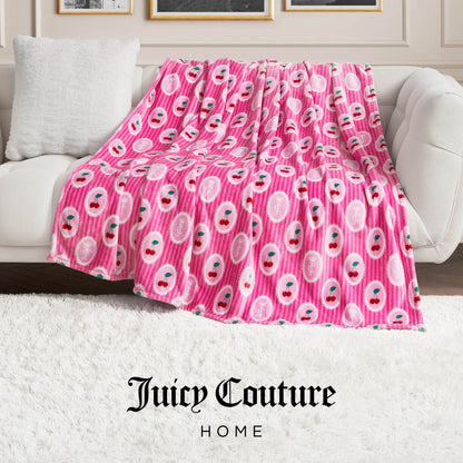 A Plush Oversized Throw Blanket in pink with cherry and Juicy Couture patterns is draped over a white sofa in a stylish living room with a white rug and elegant wall paneling. The Juicy Couture Home logo is visible at the bottom.