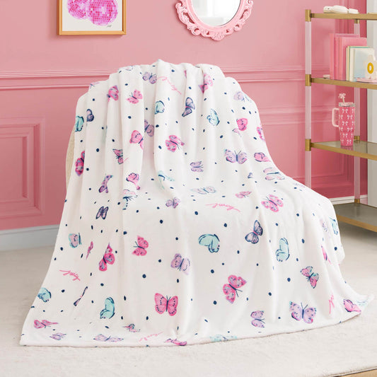 A Kids Plush Throw Blanket with colorful butterfly and polka dot patterns is draped over a chair in a pink-themed room decorated with framed wall art, a mirror, and gold shelving.