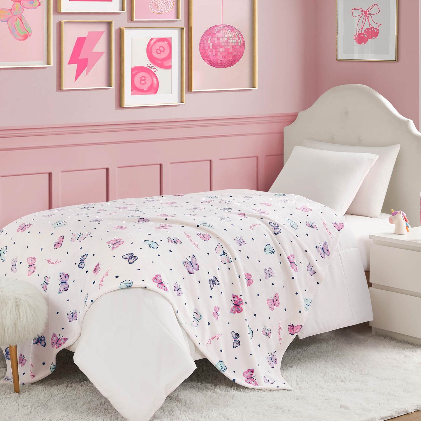 A neatly made bed with a Kids Plush Throw Blanket, ultra-soft and featuring a white and pink butterfly pattern, in a pink-themed bedroom. Pink artwork decorates the wall above the white headboard and fluffy white rug.