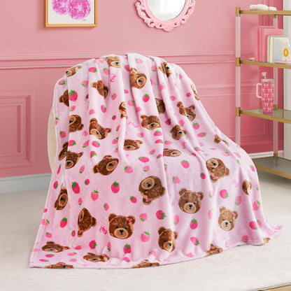 The Kids Plush Throw Blanket is draped over a beige chair in a pink, stylish room, featuring brown teddy bear faces and strawberries for a cozy, playful vibe.