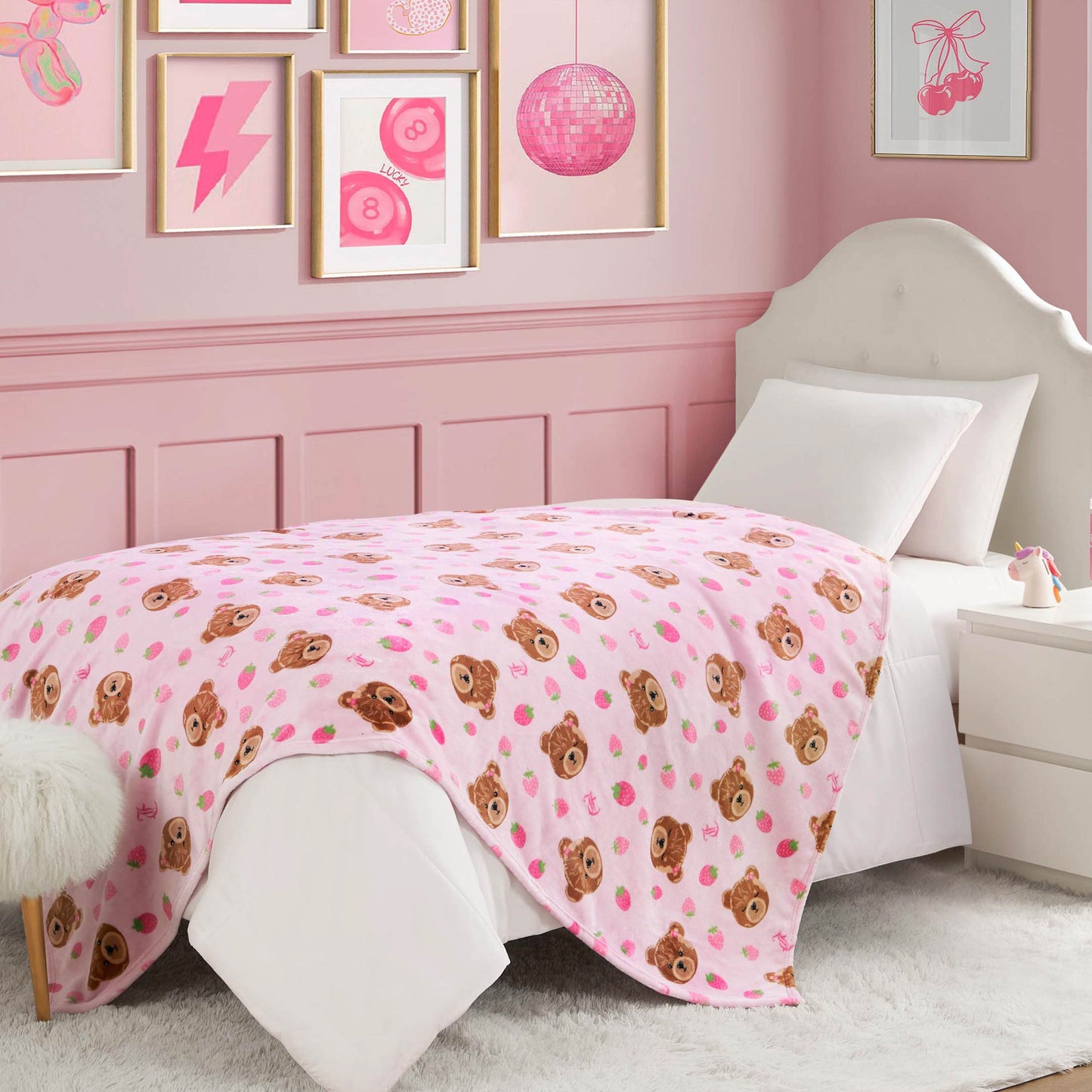 A cozy pink bedroom features a white bed topped with the Kids Plush Throw Blanket, ultra-soft and adorned with teddy bear faces. Framed pink art decorates the walls, and a nightstand sits beside the bed for extra charm.