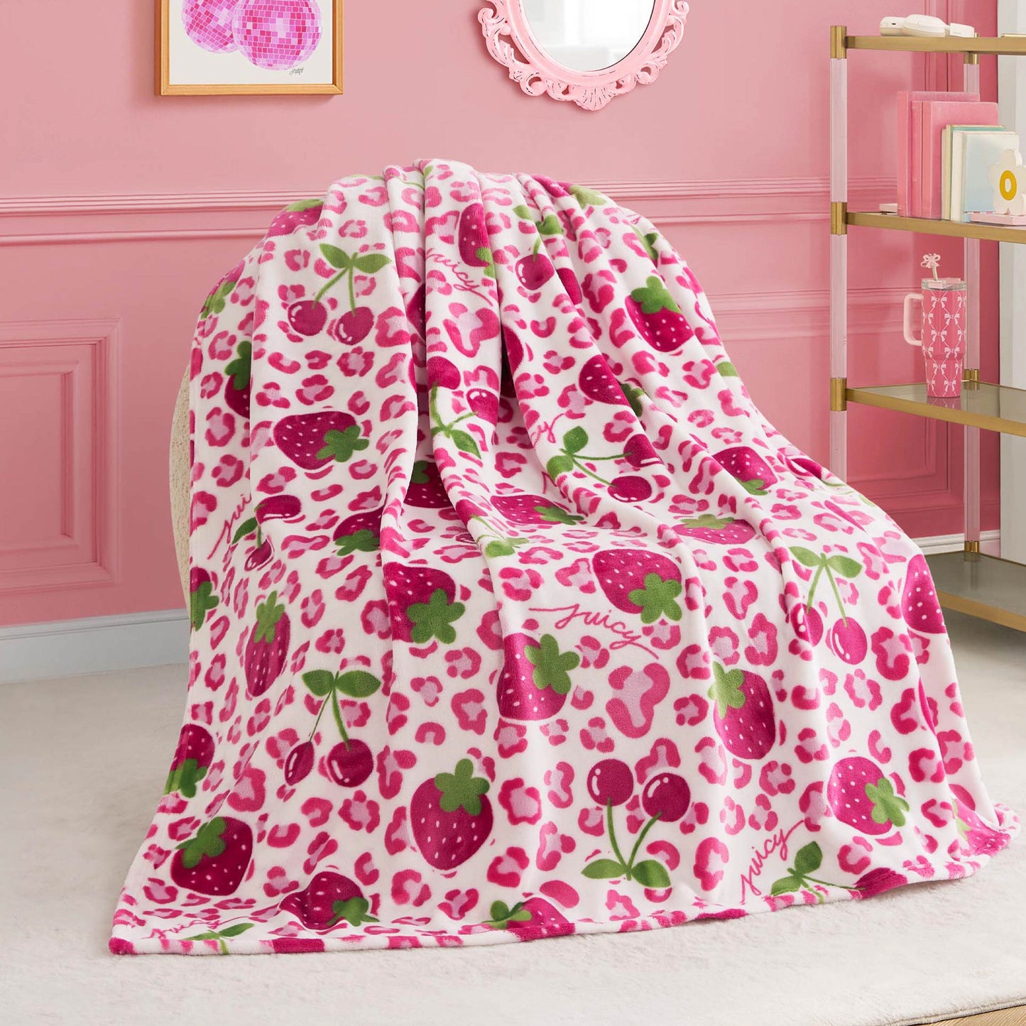 A Kids Plush Throw Blanket with pink leopard spots, strawberries, cherries, and green leaves is draped over a chair in a pink room with gold shelves and framed artwork on the wall.